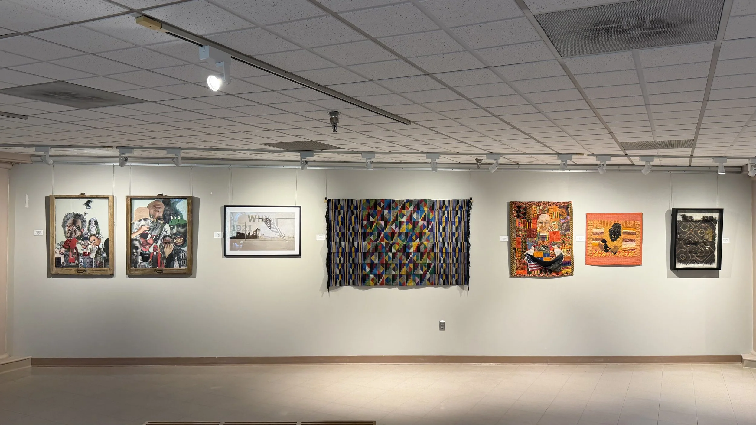 An art gallery showcasing multiple colorful paintings and textile art on a white wall, with ceiling lights directed towards the artwork.