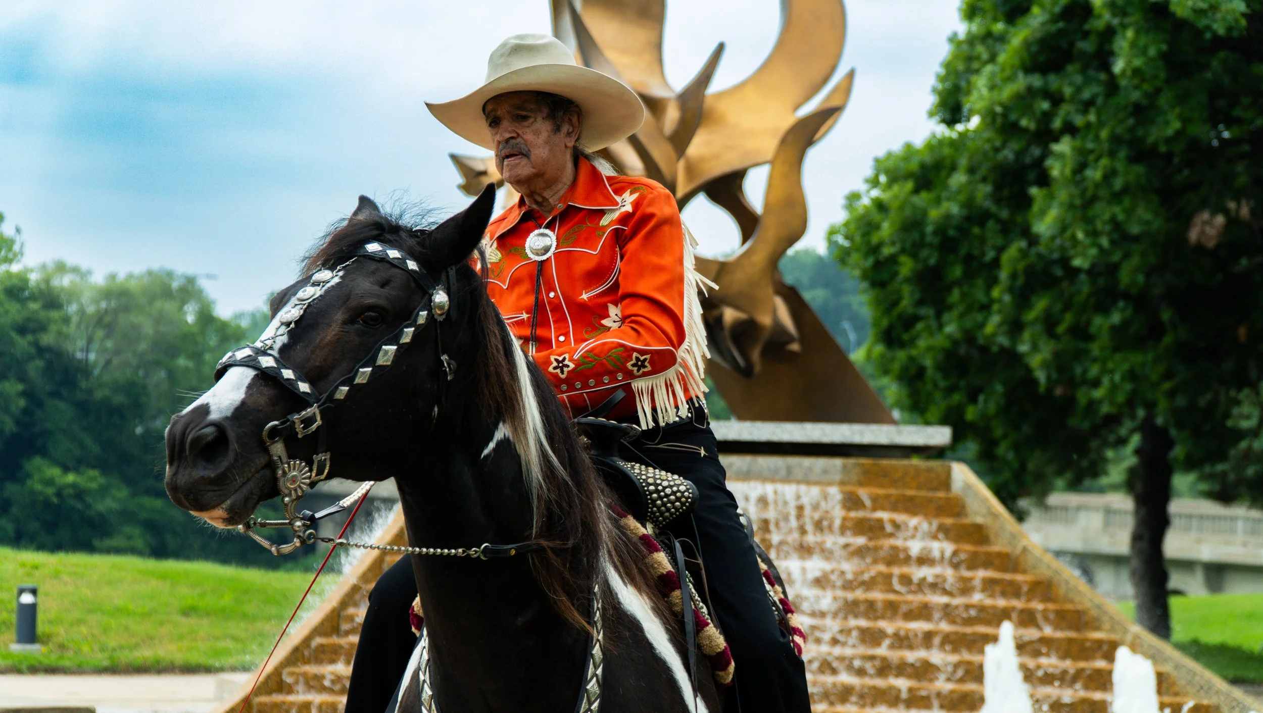Rex Purefoy wearing a cowboy hat and an orange Western shirt with embroidery, riding a decorated black and white horse. In the background, there is a sculpture with large gold-colored abstract shapes and greenery with trees.