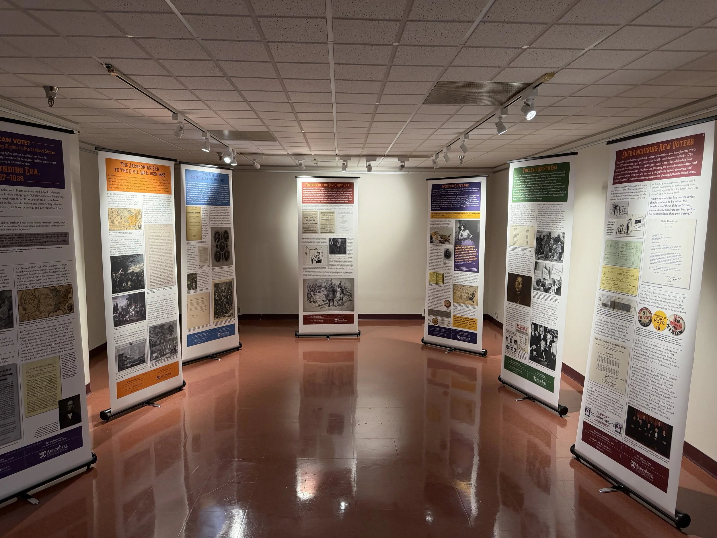 An exhibition displaying multiple informational posters on stands, arranged in a semi-circle in a room with a tiled ceiling and reflective floor.
