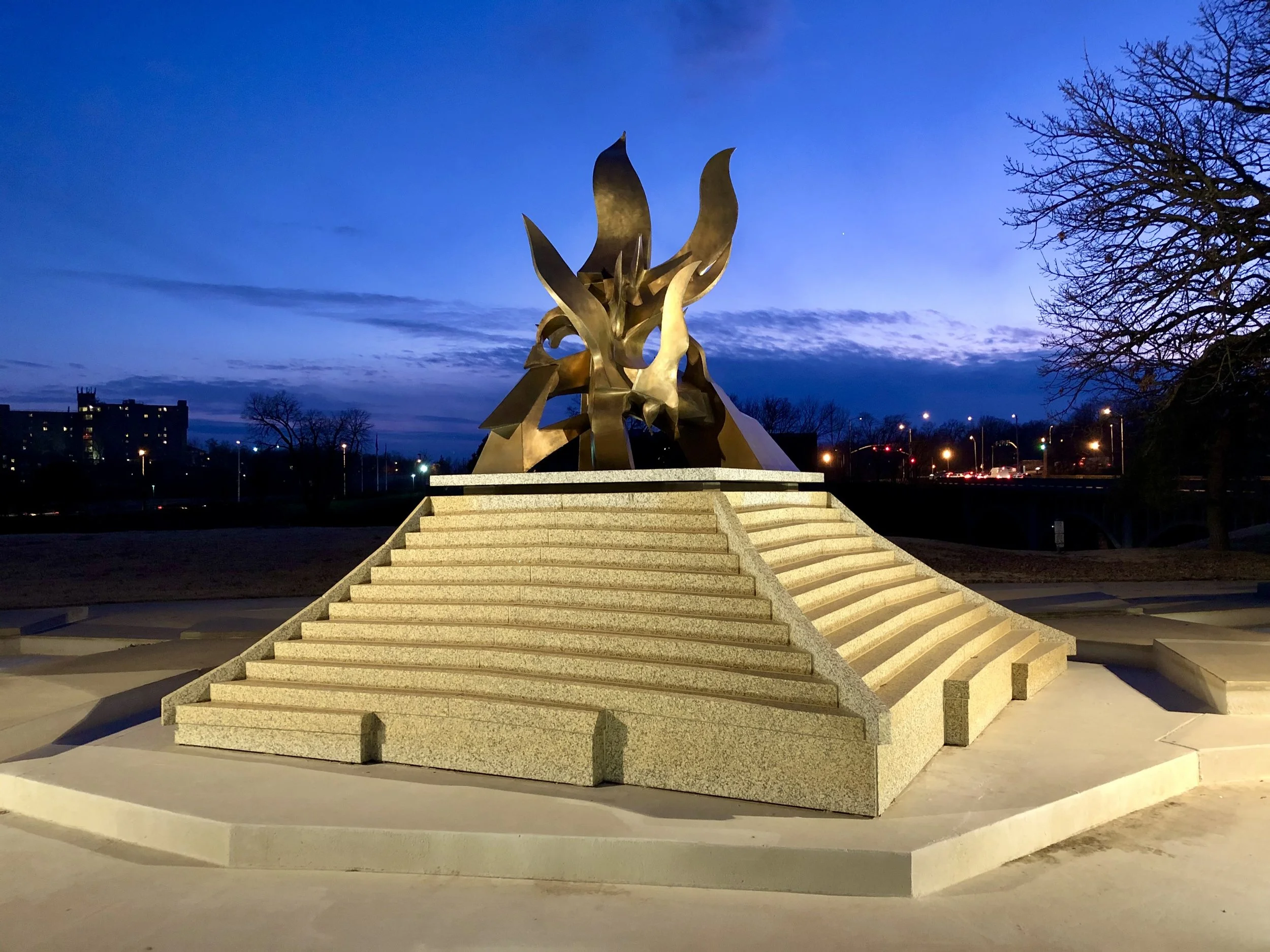 A large, modern sculpture resembling flames, made of golden metal, is situated on a raised platform with steps. It is set against a twilight sky with some clouds and city lights in the background, and there's a leafless tree on the right side.