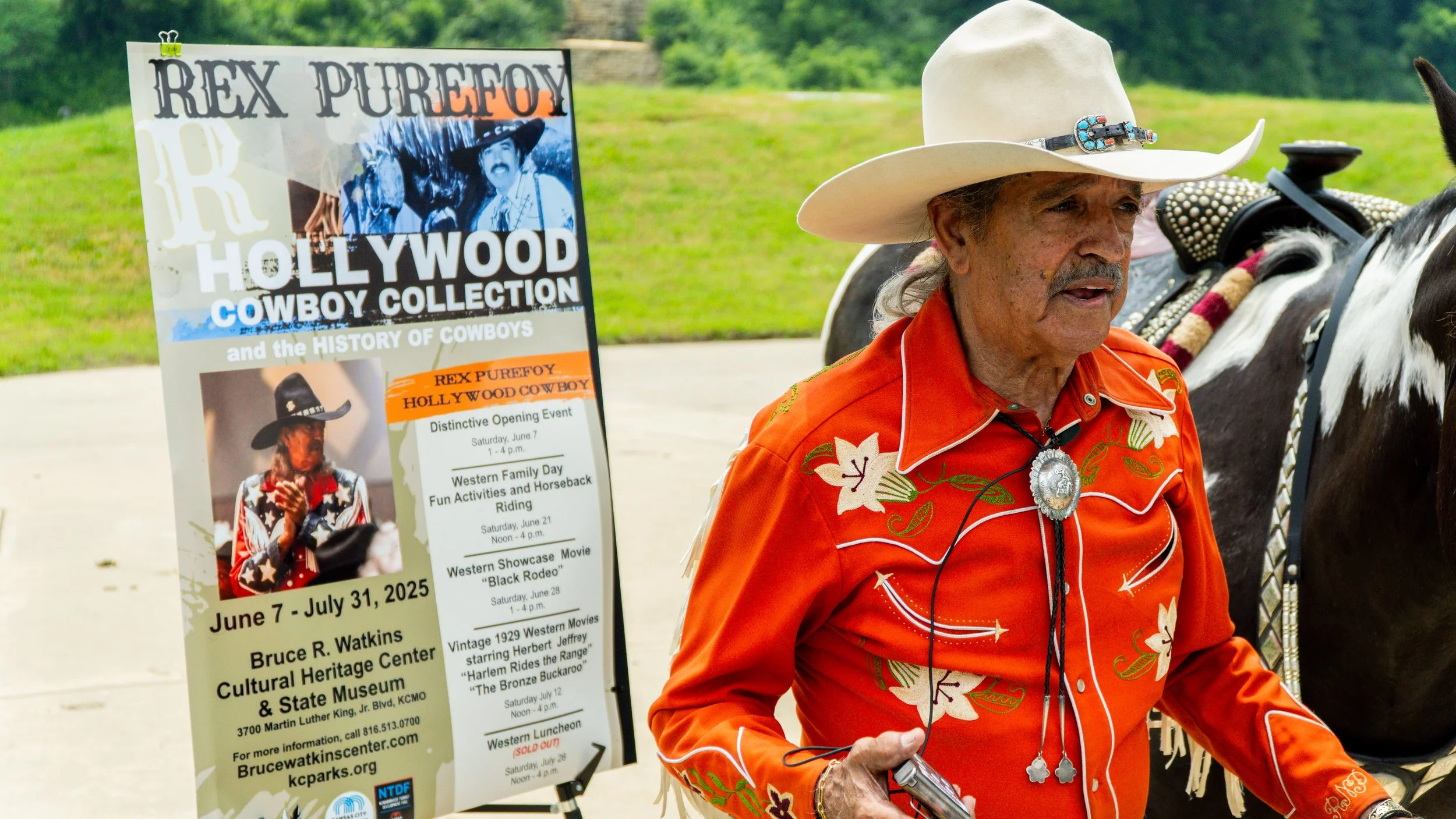 Rex Purefoy dressed in a bright orange Western shirt with floral embroidery and pearl accents, wearing a star-shaped brooch, white cowboy hat with a turquoise and silver buckle, walking outdoors with a horse.