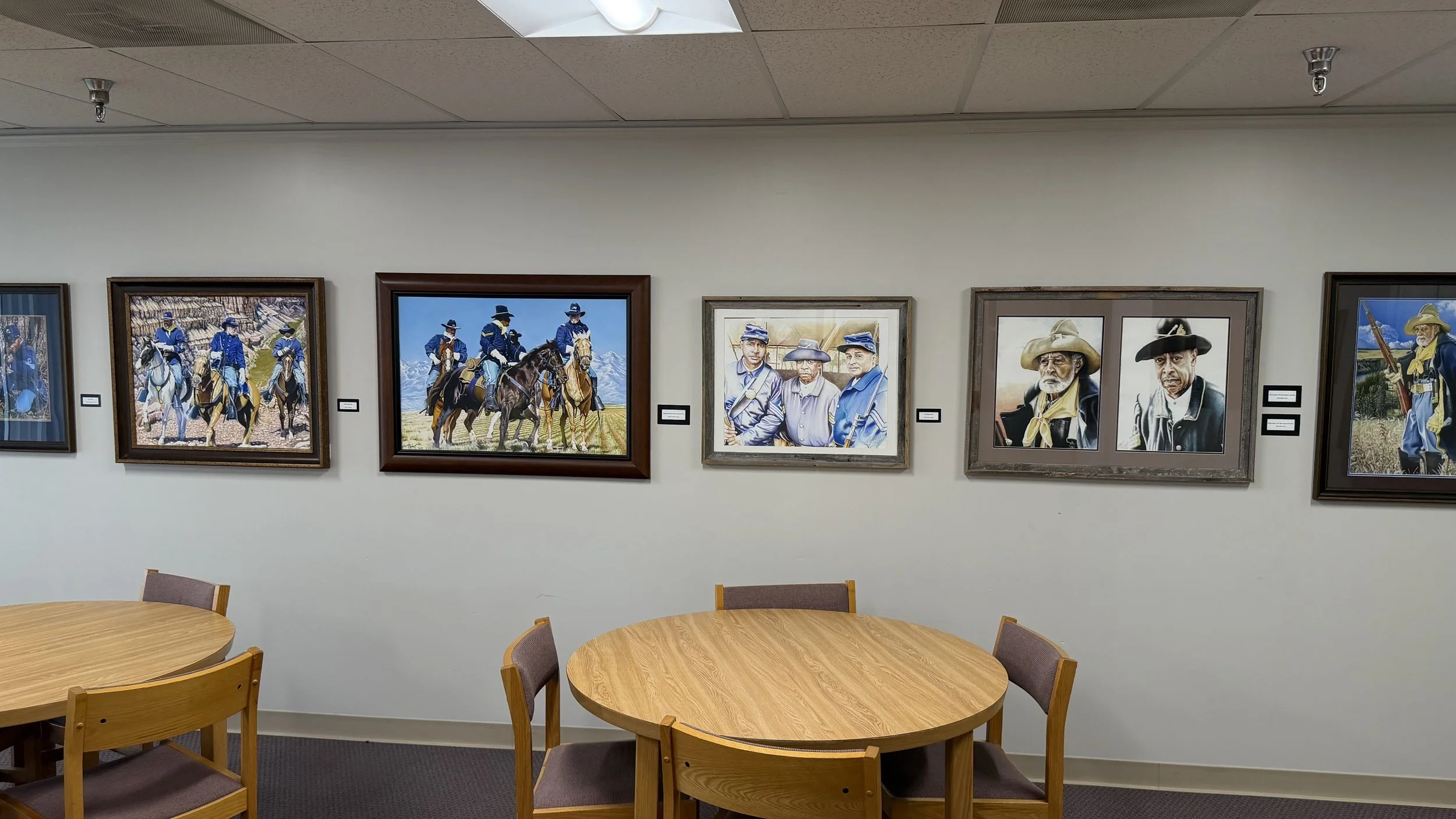Gallery wall with paintings of Buffalo Soldiers and scenes, with wooden tables and chairs in front.