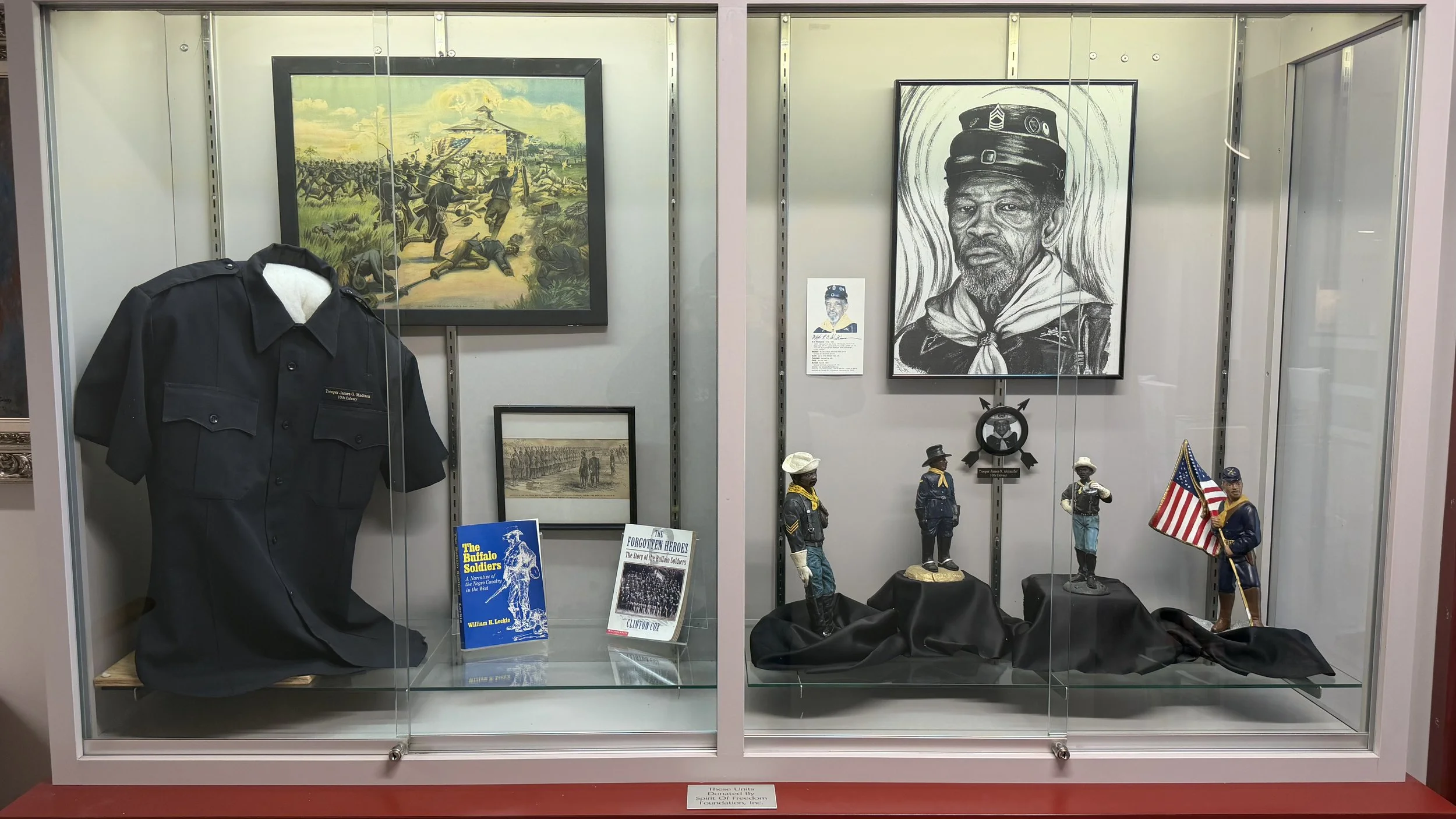 Historical display case featuring Buffalo Soldiers artifacts including a uniform, figurines of soldiers, a large black-and-white portrait of a man in a military hat, various paintings depicting battles about historical soldiers.