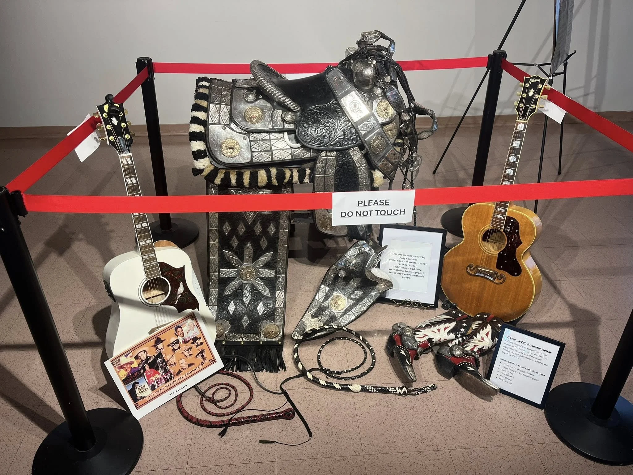 Display of vintage and modern guitars, a saddle with decorative metalwork, jewelry, and posters, all enclosed by a red velvet rope at a museum exhibit.