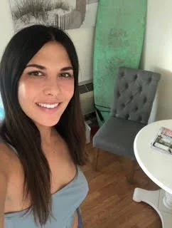 A woman with long dark hair smiling in a room with a green surfboard, a gray tufted chair, and a round white table.