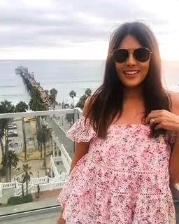 A woman with long dark hair wearing sunglasses and a pink off-shoulder floral dress, smiling on a balcony with a view of the ocean and a pier in the background.
