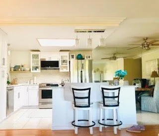 Bright kitchen with white cabinets, a kitchen island with two bar stools, and a living room area in the background.