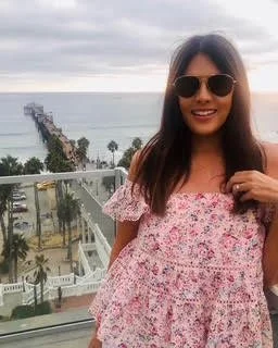 A woman wearing sunglasses and a pink floral off-shoulder dress, standing outdoors with a pier, ocean, and palm trees in the background.