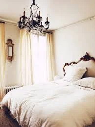 A cozy bedroom with a bed, a chandelier hanging from the ceiling, a window with curtains, and a wall clock.