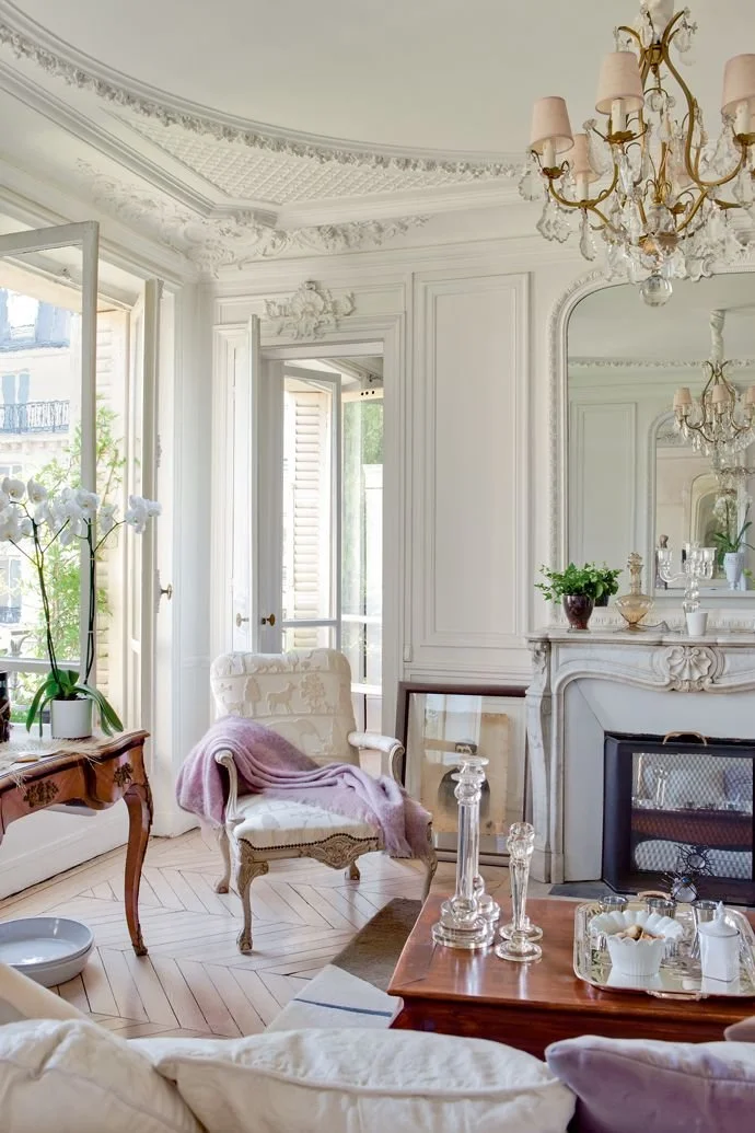 Elegant living room with white walls and crown molding, large windows with open shutters, crystal chandeliers, a white fireplace with decorative items, a white armchair with a pink blanket, glass decanters and a tray on a wooden table, a wooden side table with orchids, and a mirror reflecting the room.