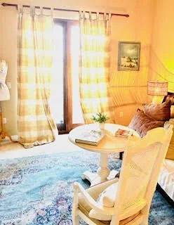 Cozy living room with a window, checkered curtains, a small round table with a book and plant, a comfy sofa with pillows, a lamp, and a framed picture on the wall.