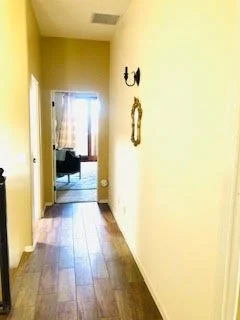 Empty hallway with light yellow walls, wooden flooring, and a small decorative mirror hanging on the right wall; a black wall-mounted light fixture is visible, with a view into a room at the end of the hallway.