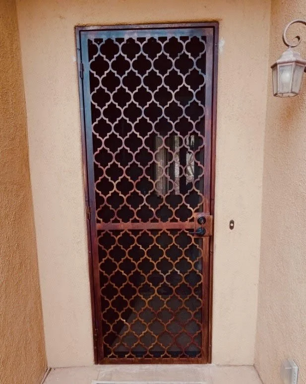 A metal security door with a geometric pattern, next to a wall-mounted outdoor lantern light.
