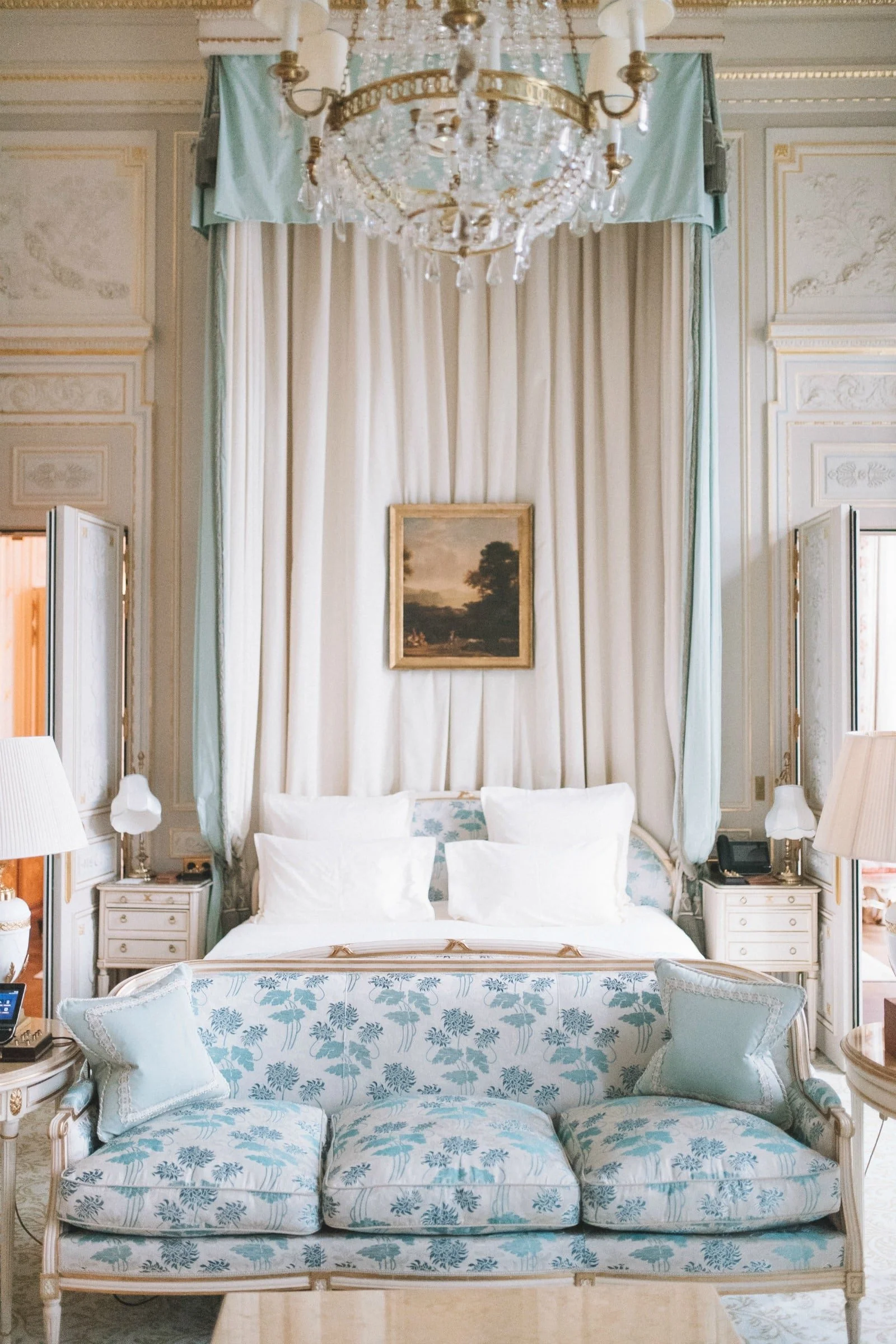Elegant bedroom with a canopy bed, light blue floral upholstery, and matching sofa. Cream and gold accents, classic artwork, and a crystal chandelier.