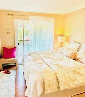 A cozy bedroom with a large bed covered in cream-colored bedding, a wooden nightstand with a bright pink pillow, a tall lamp, and a sliding glass door with white curtains leading to a balcony.