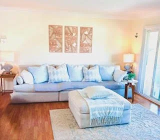 Living room with a large light gray sofa, white cushions, a matching ottoman, two side tables with lamps, wall art above the sofa, hardwood flooring, and a sliding glass door with curtains.