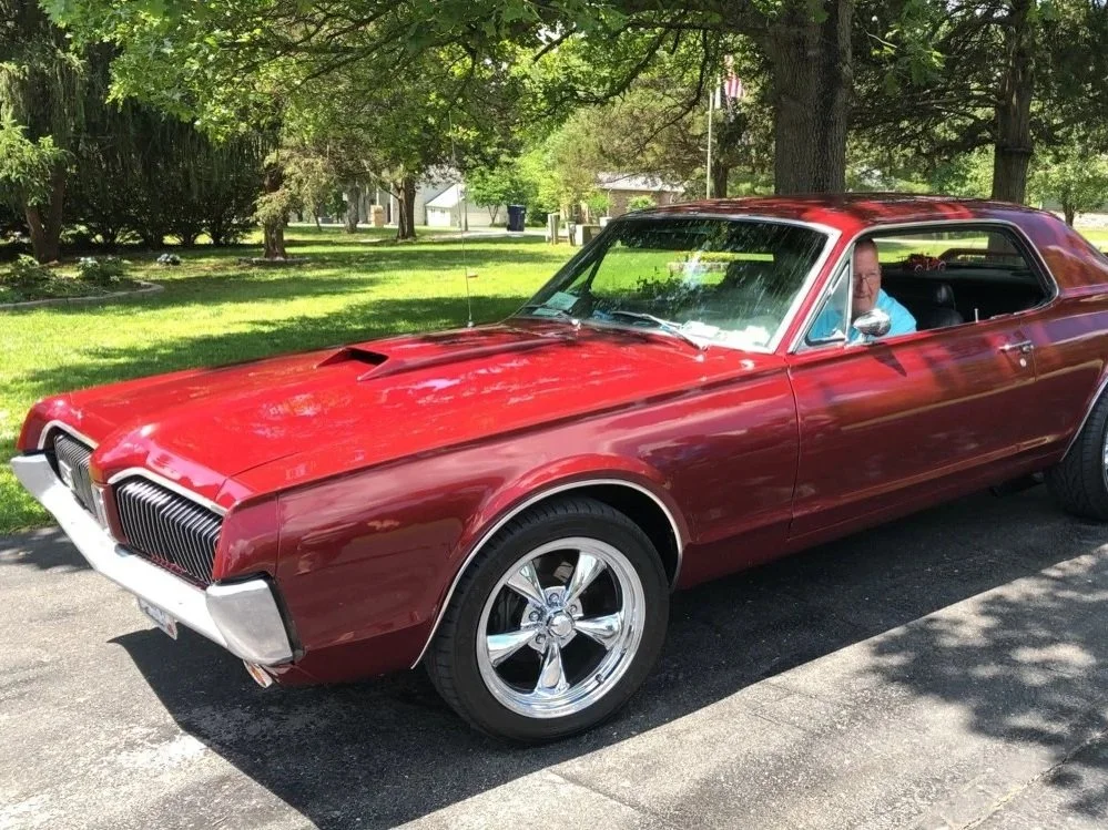 Red vintage car parked on driveway with a man sitting inside, shaded by trees under a bright sky.