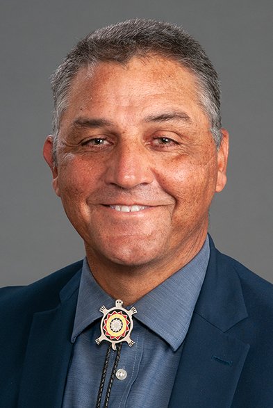 A photo of Ronny Bell (Lumbee), PhD, MS, in a blue suit with a tribal badge on his shirt collar, smiling at the camera.