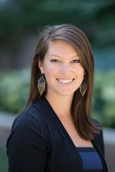 A photo of Stephanie Wheeler, PhD MPH with brown hair smiling outdoors, wearing earrings and a dark jacket.
