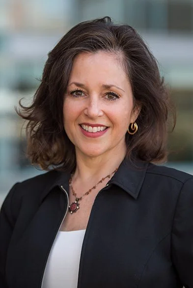 A photo of Barbara Alvarez Martin, DrPH MPH, with shoulder-length wavy brown hair, smiling, wearing gold hoop earrings, a dark blazer, and a necklace, standing outdoors with a blurred building background.