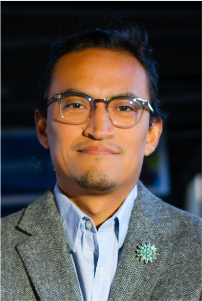 A photo of Marc Emerson (Navajo/ Diné), PhD, wearing glasses, a gray blazer, and a light blue shirt, smiling at the camera.