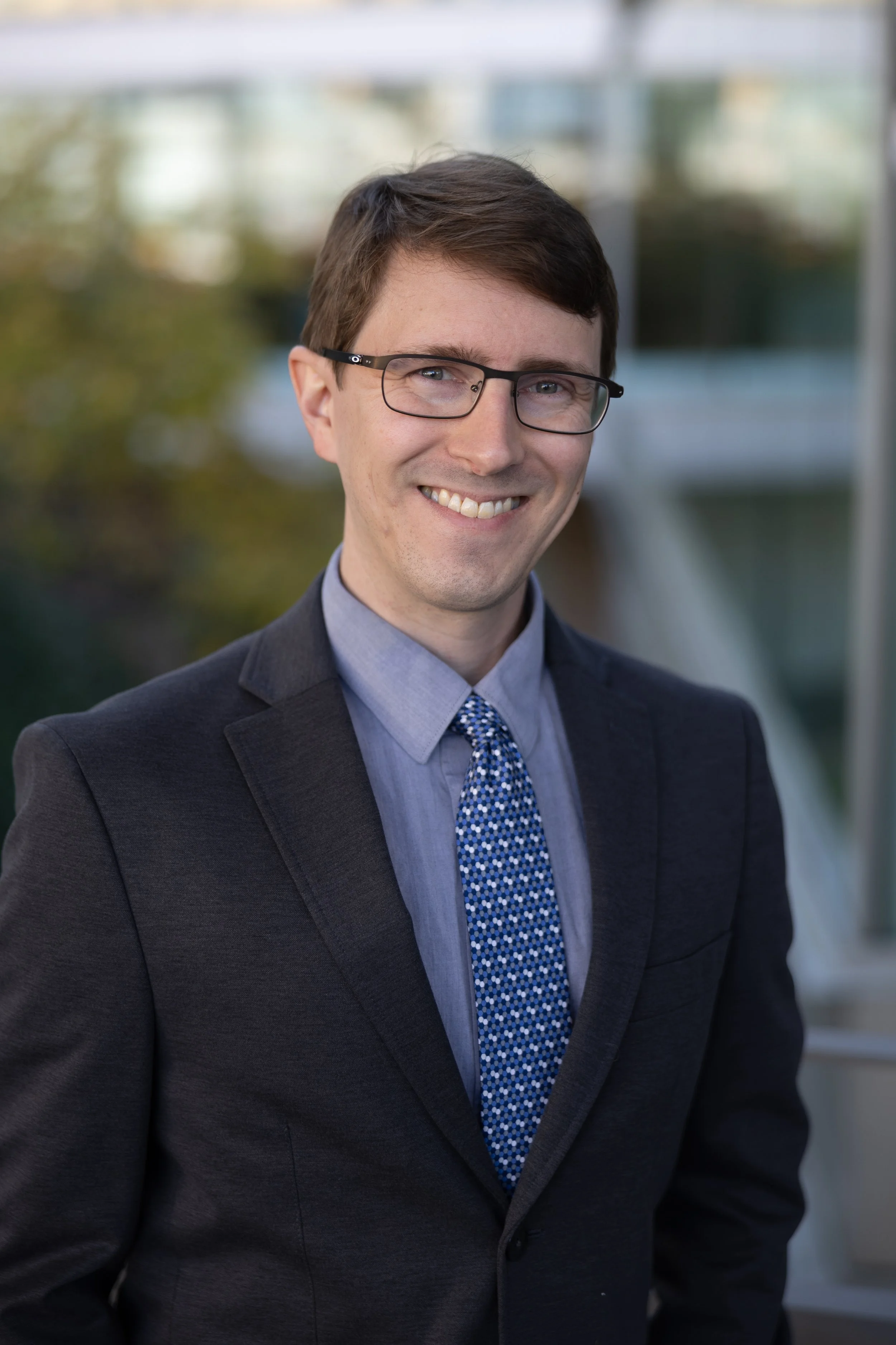 A photo of Andrew Dodgson, DPT, with short brown hair, wearing glasses, a dark suit, blue shirt, and patterned blue tie, smiling with a blurred cityscape background.