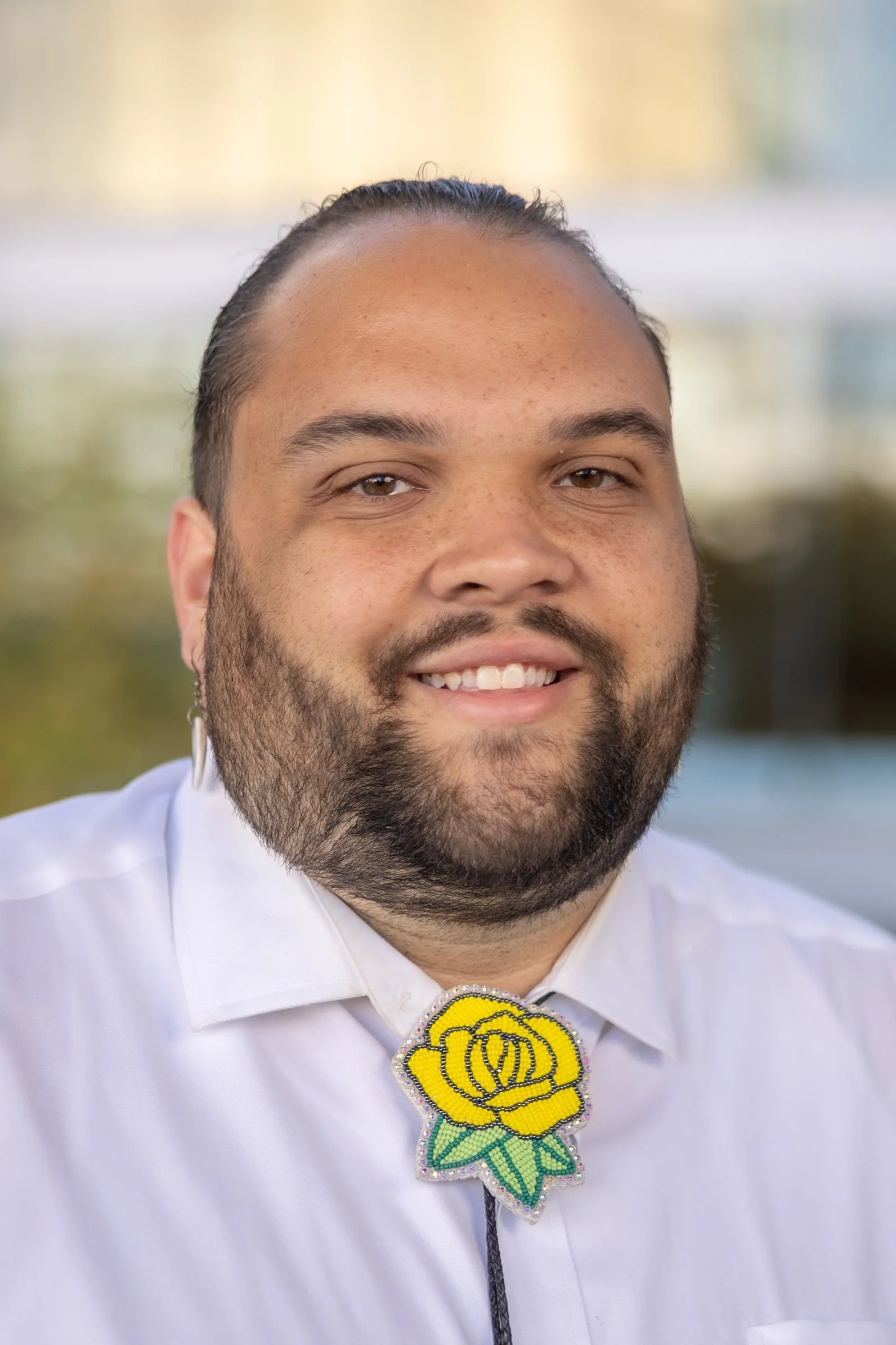 A photo of Ryan Dial (Lumbee), MCLS MLS (ASCP) CM, with short dark hair, a beard, and light brown eyes wearing a white shirt and a yellow beaded brooch with a green leaf detail on his collar, outdoors with a blurred background.