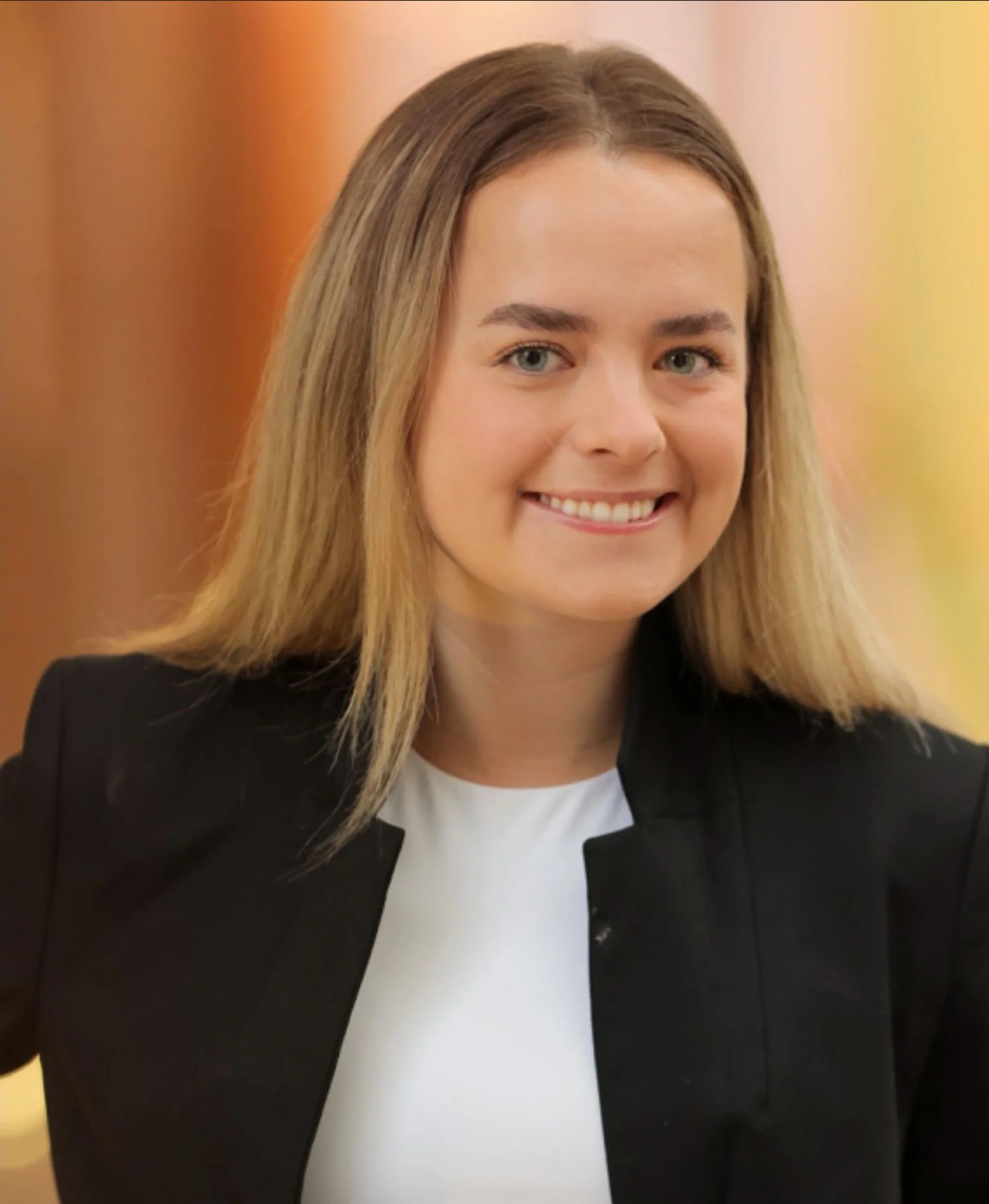A photo of Erica Fennimore (Tlingit) with shoulder-length blonde hair, blue eyes, and a bright smile, wearing a black blazer over a white shirt, standing in front of a blurred warm-toned background.
