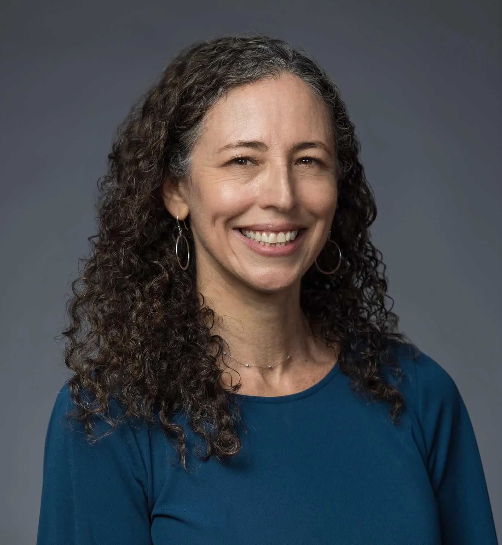A photo of Meira Epplein, PhD, MS, MA, with curly dark hair, wearing earrings, a necklace, and a blue shirt, against a plain gray background.