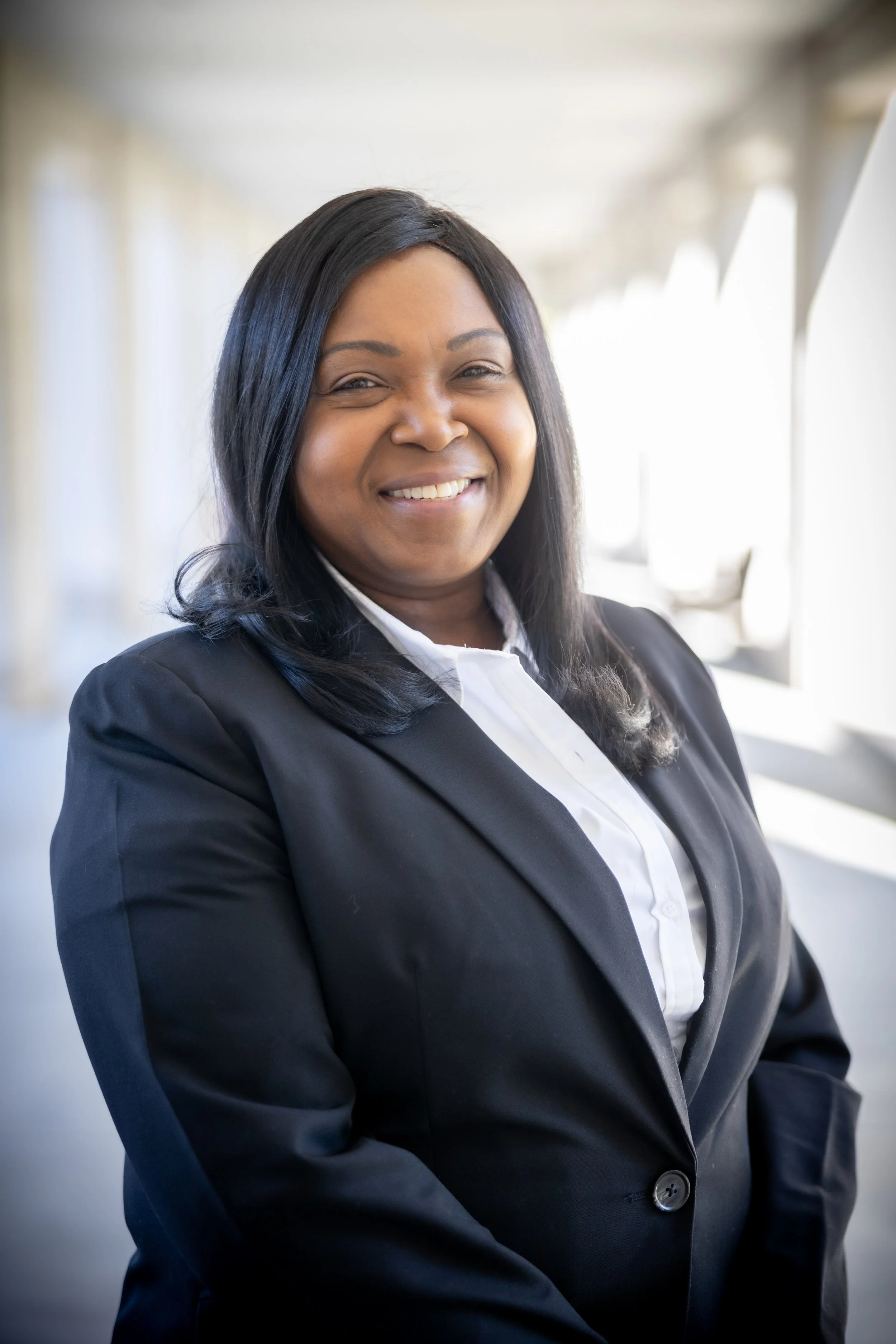 A photo of LaSonia Barnett, MA, with long black hair and a friendly smile, dressed in a black blazer and white shirt, standing in front of large windows in an office setting.