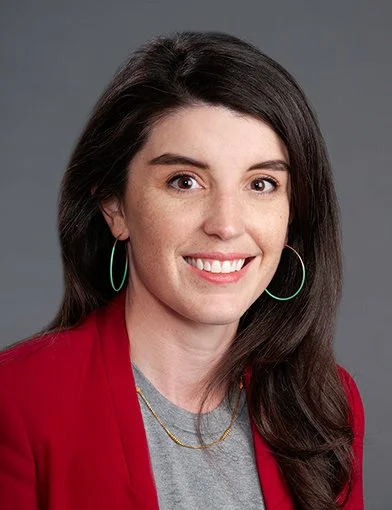 A photo of Rachel Denlinger-Apte, PhD MPH, with long dark hair smiling, wearing hoop earrings, a gray t-shirt, and a red blazer against a gray background.