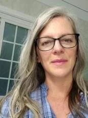 A photo of Sydnee Crankshaw, MPA, with long gray hair and glasses outside near a building with white window frames.