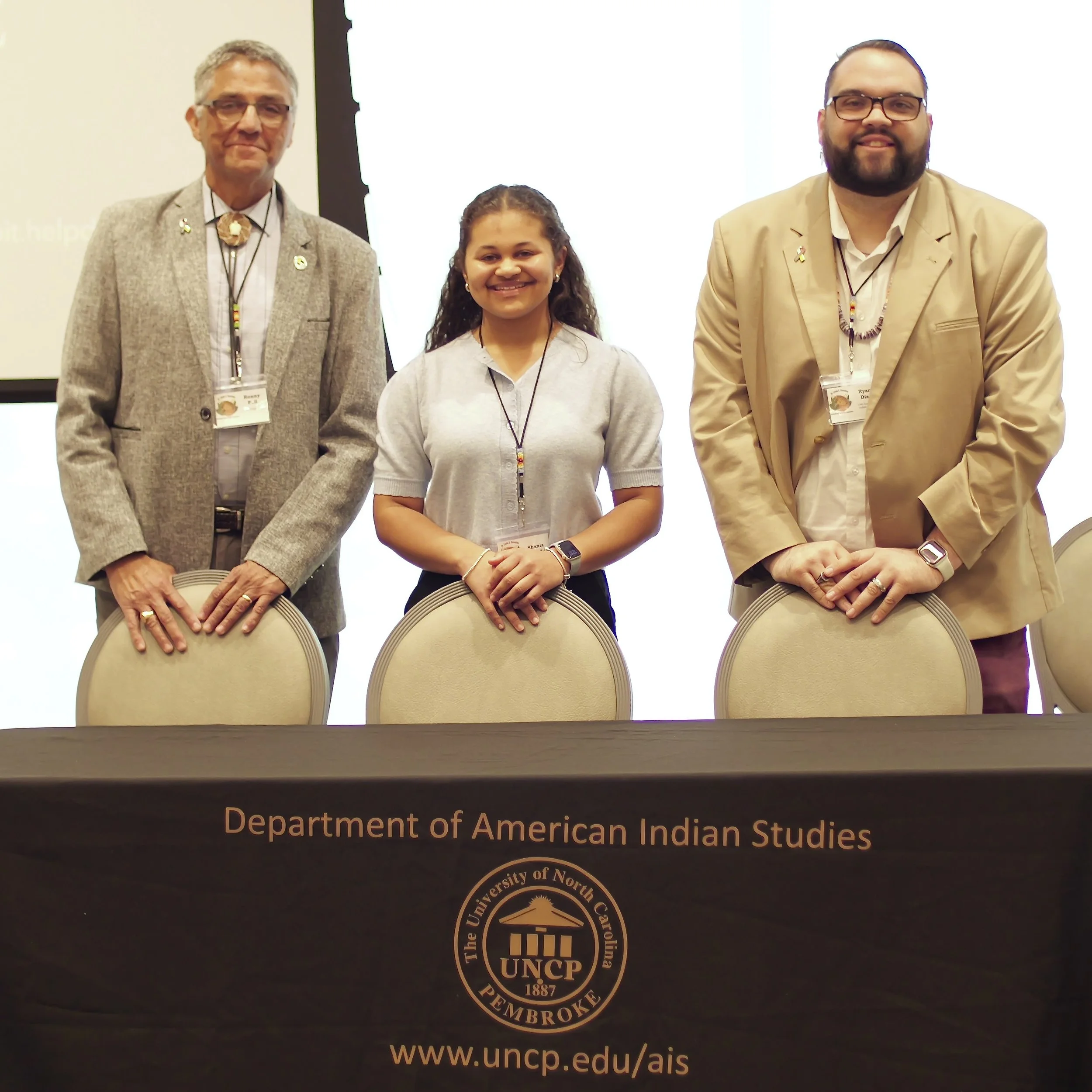 Three individuals standing behind chairs at a conference or event, with a presentation screen in the background. They are wearing formal attire with name tags and appear to be posing for a group photo. The bottom of the image shows part of a logo for the Department of American Indian Studies at the University of North Carolina Pembroke.