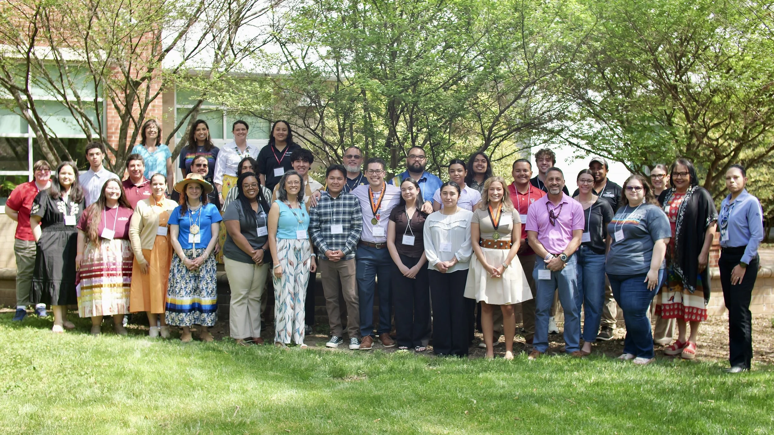 Group of diverse people outdoors, smiling, posing for a photo in front of green trees and a brick building.