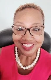 A photo of Gwendolyn Shaw, smiling, wearing glasses, a pink top, and a pearl necklace, sitting in an office chair.