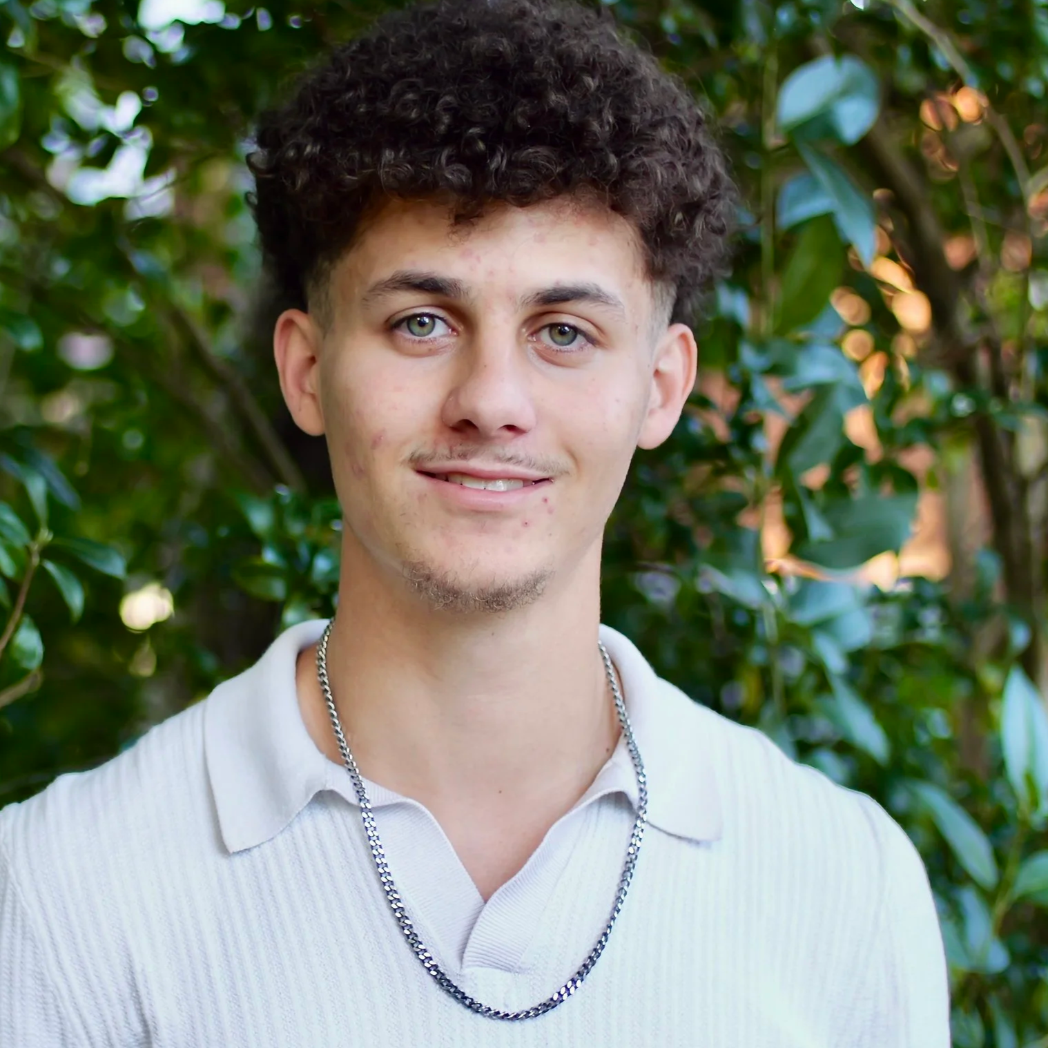 A photo of Cayden Hammonds (Lumbee) with curly hair, light skin, and green eyes, wearing a white polo shirt and silver chain necklace, standing in front of leafy green bushes.