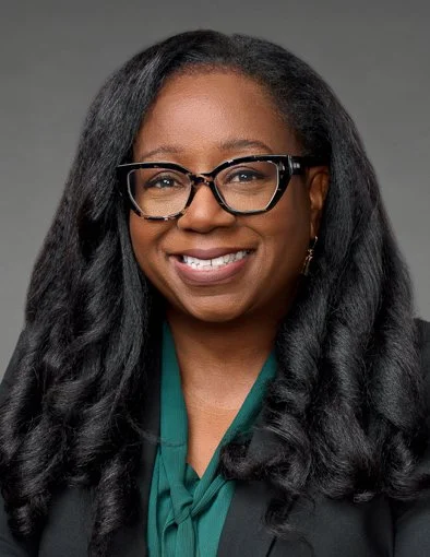 A photo of Nadine Barrett, PhD, MS, MPH, with long black hair, wearing glasses and a dark blazer, smiling at the camera against a neutral background.