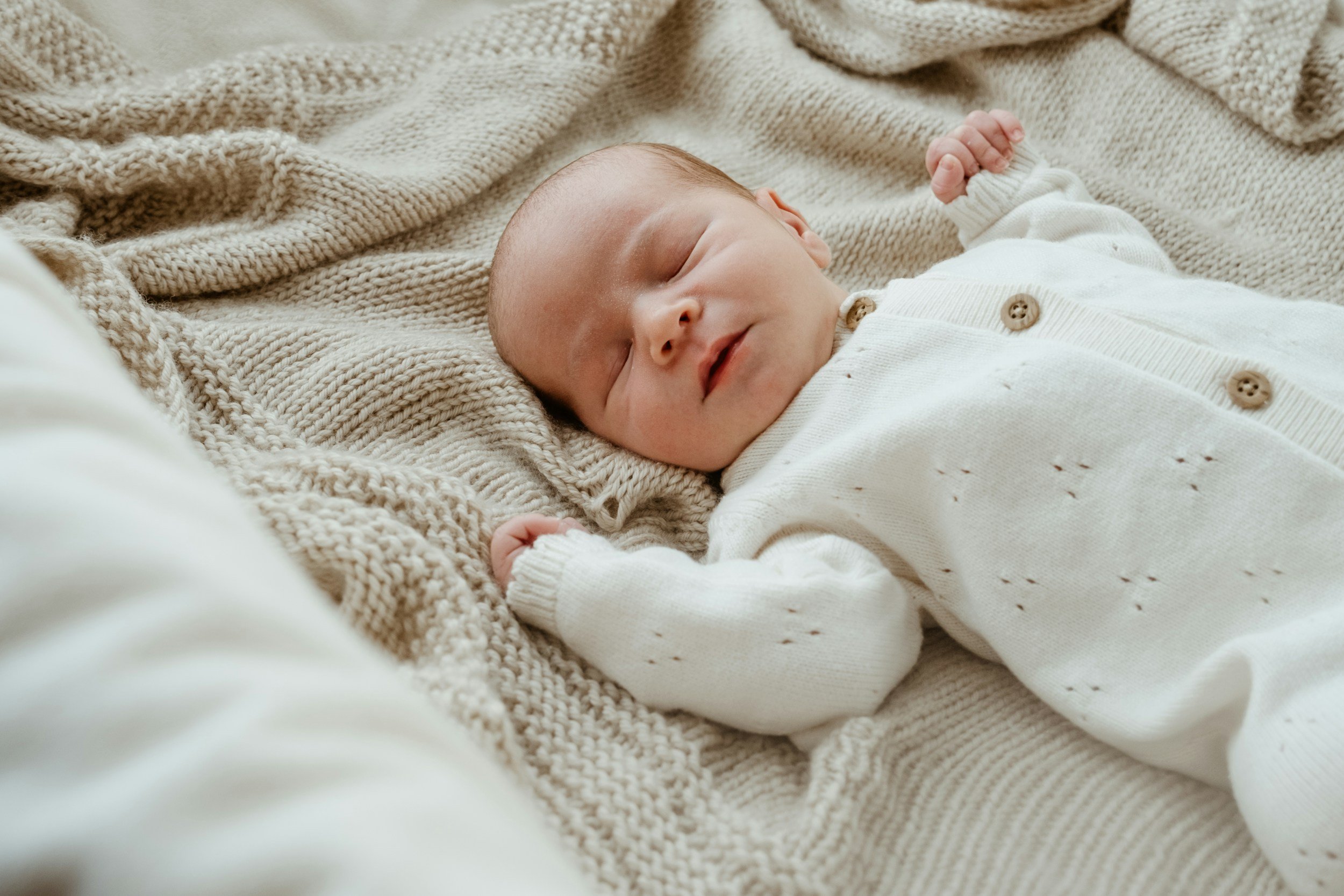 A sleeping baby lying on a soft, knitted cream-colored blanket, wearing a cream-colored cardigan with wooden buttons.