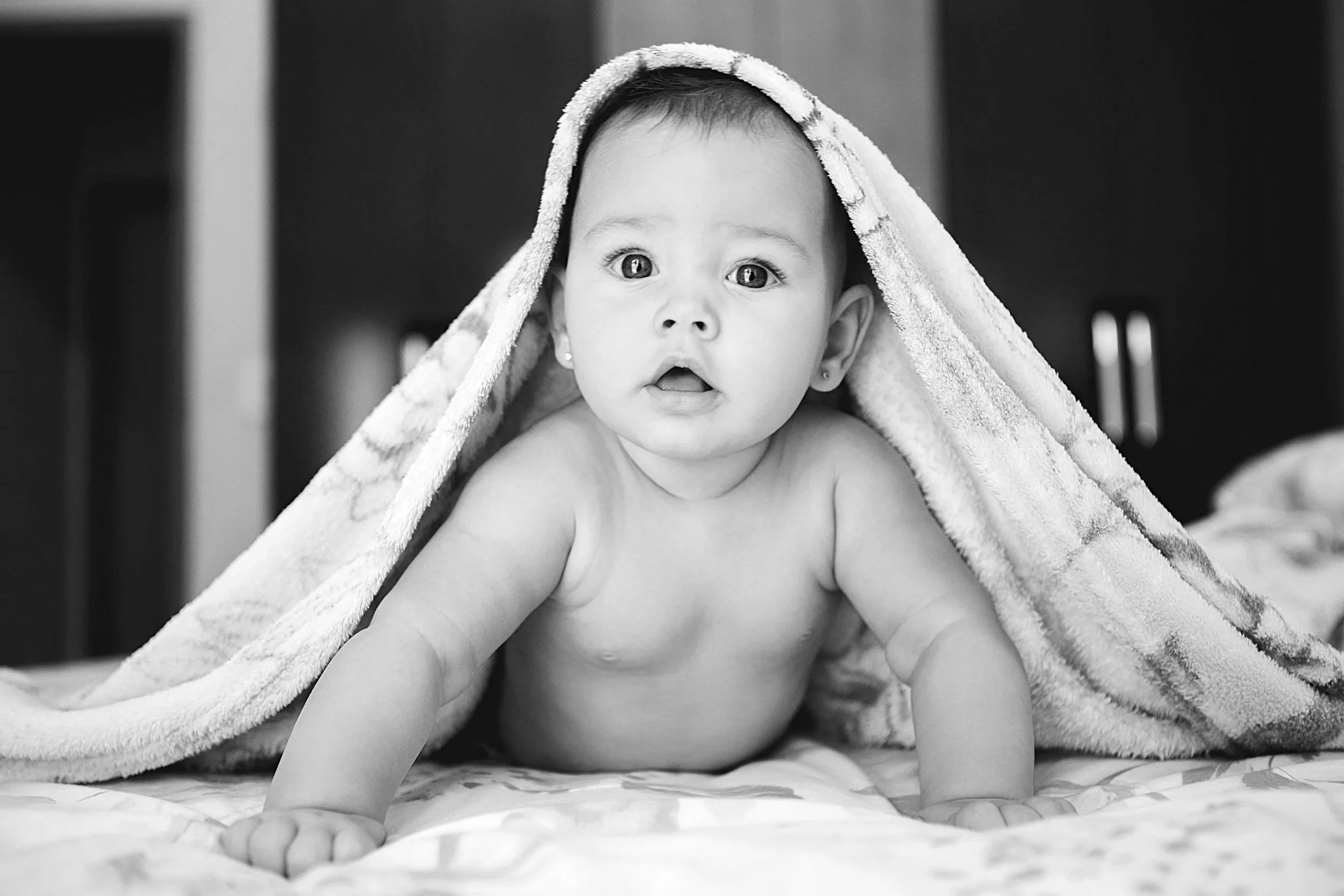 A black-and-white photograph of a baby with wide eyes, lying on a blanket, partially covered with a towel over the head, looking directly at the camera.