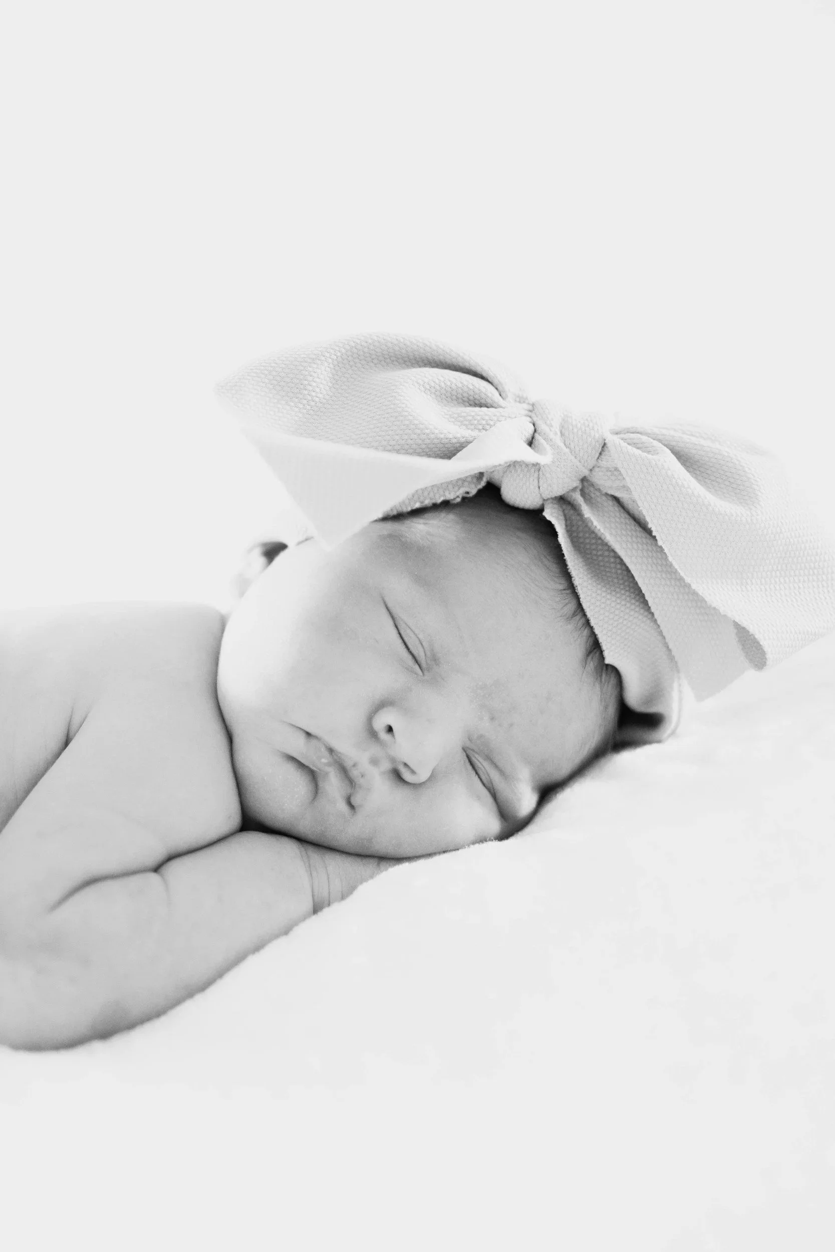 Black and white photo of a sleeping baby with a bow headband, lying on a soft surface.