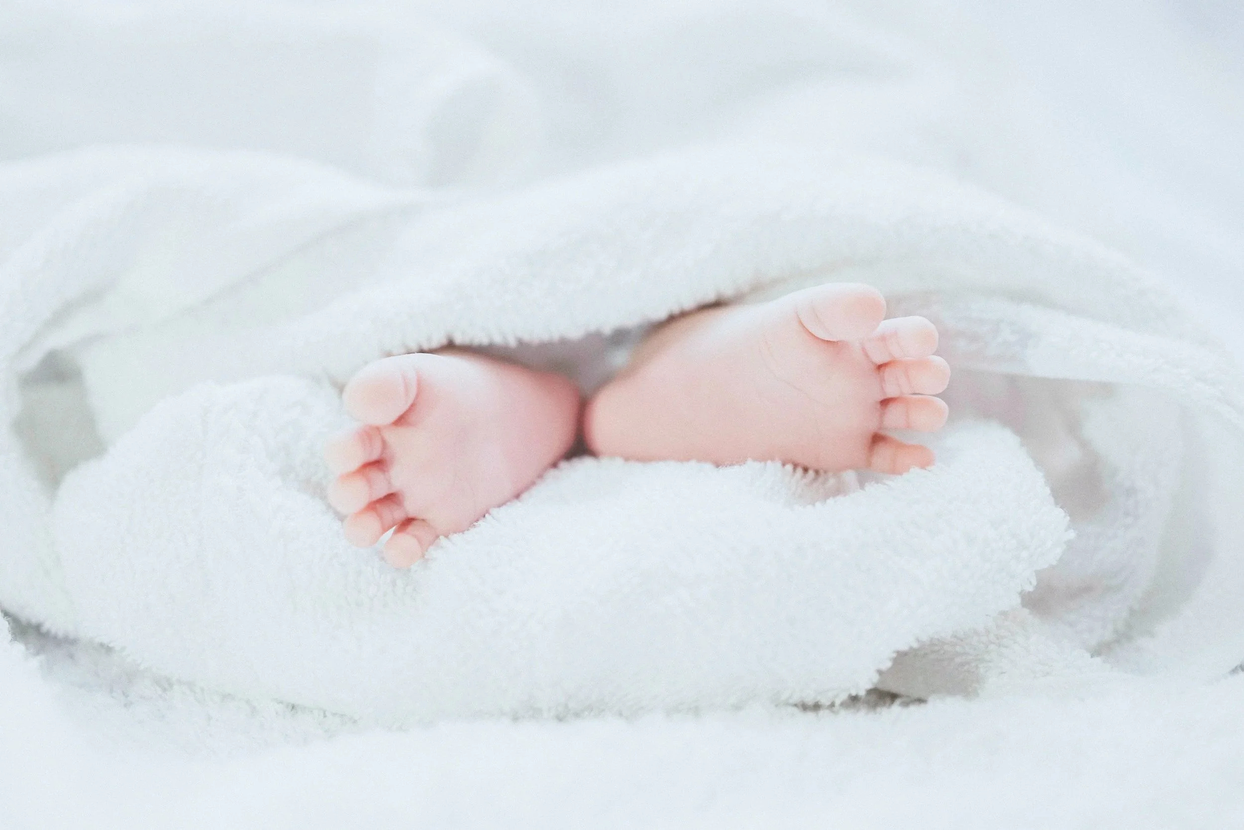 Close-up of a baby's feet peeking out from under a soft, white blanket on a white, fluffy surface.