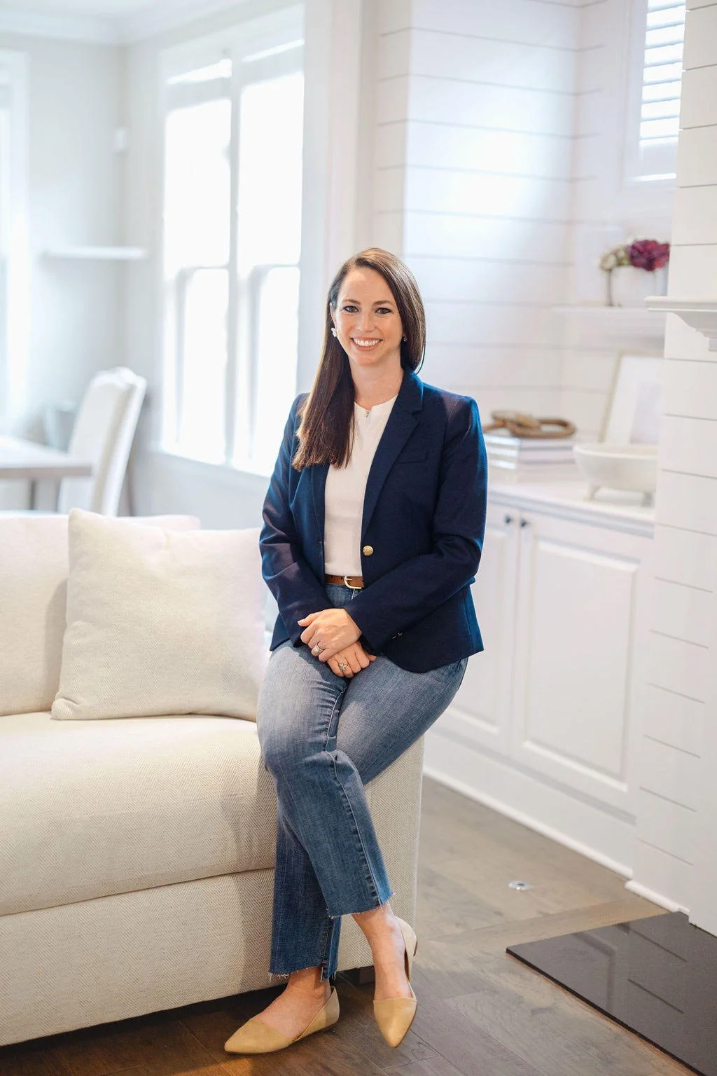 A woman with long brown hair smiling, sitting on a beige couch in a bright, white room with windows, white walls, and minimal decor, wearing a navy blazer, white top, gray jeans, and beige heels.