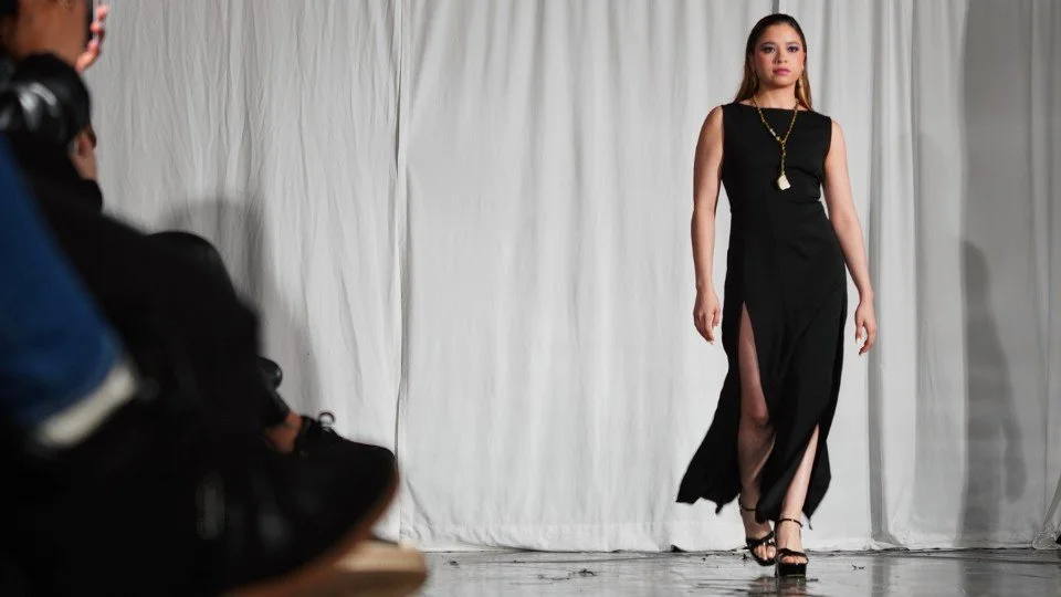 A woman in a black dress with high slits walking on a runway, with a person in dark clothing taking a photo or recording nearby.