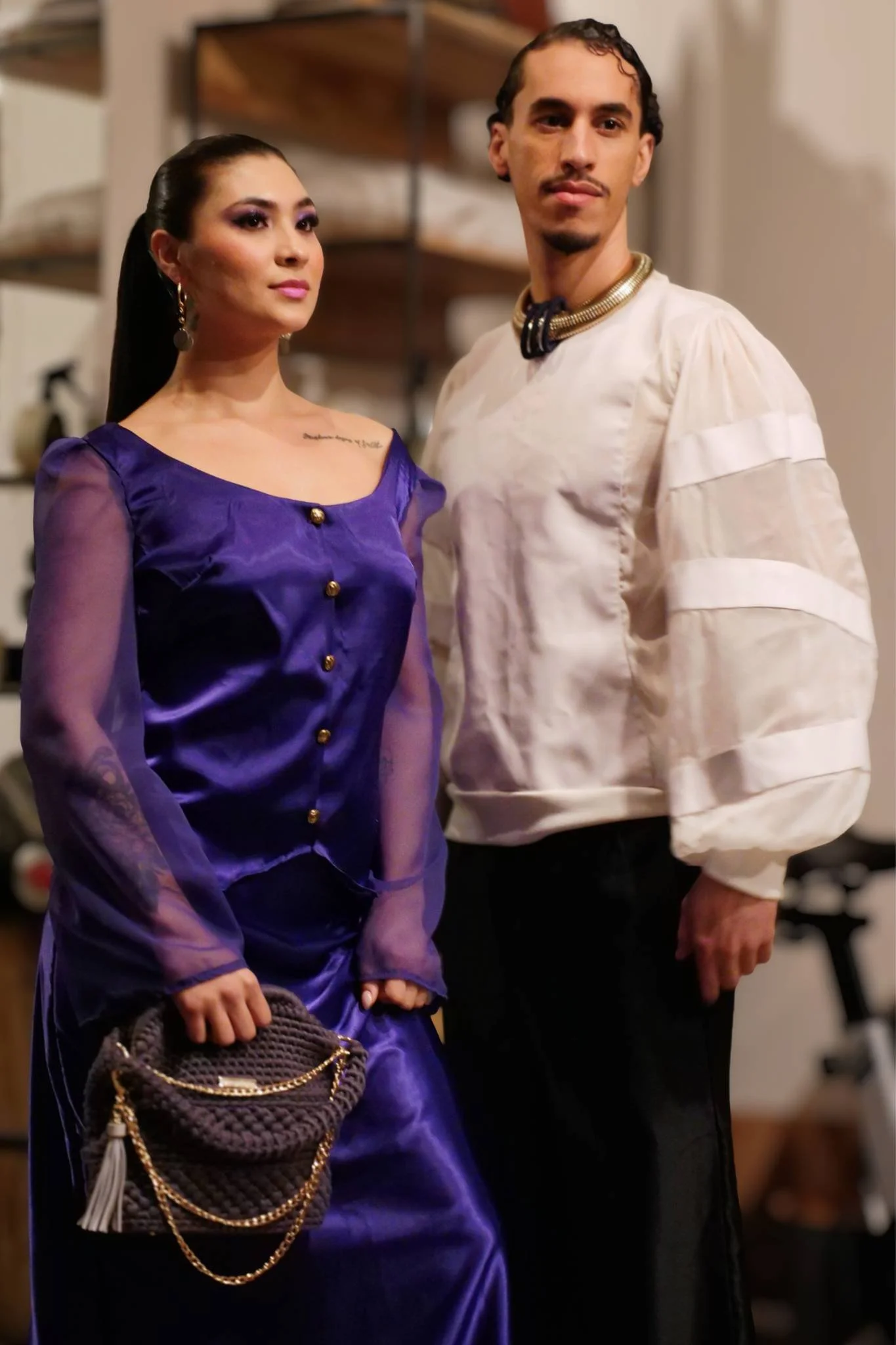 A woman with dark hair in a purple satin dress holding a small knitted brown purse with gold chains and a tassel, standing next to a man in a cream-colored shirt with a gold necklace, in an indoor setting.