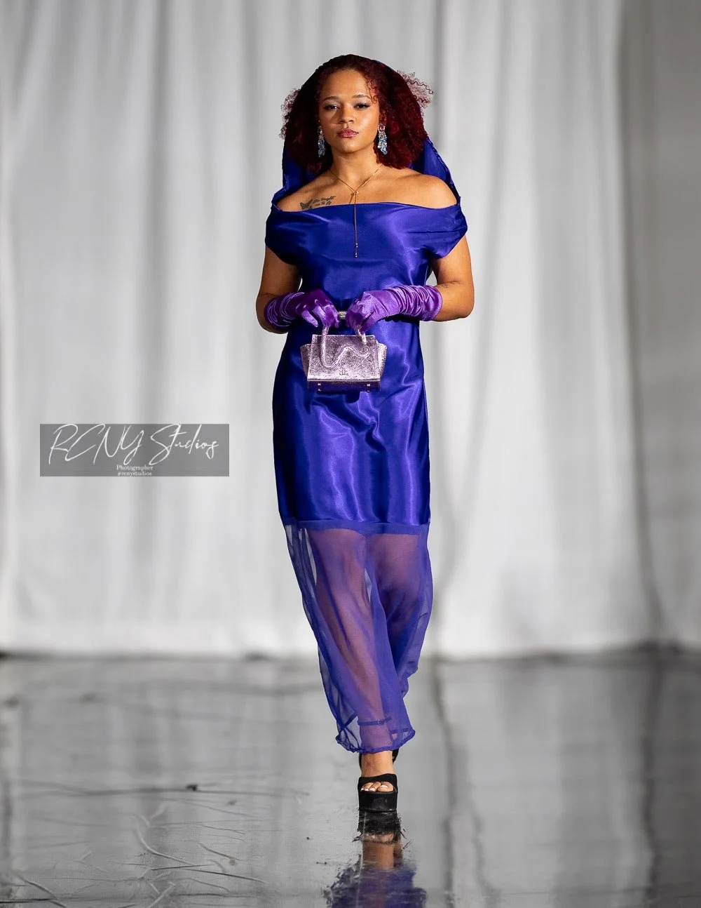 A woman walking on a runway in a shiny blue dress with a sheer bottom, wearing purple gloves, accessorized with earrings, a necklace, and carrying a small purple handbag.