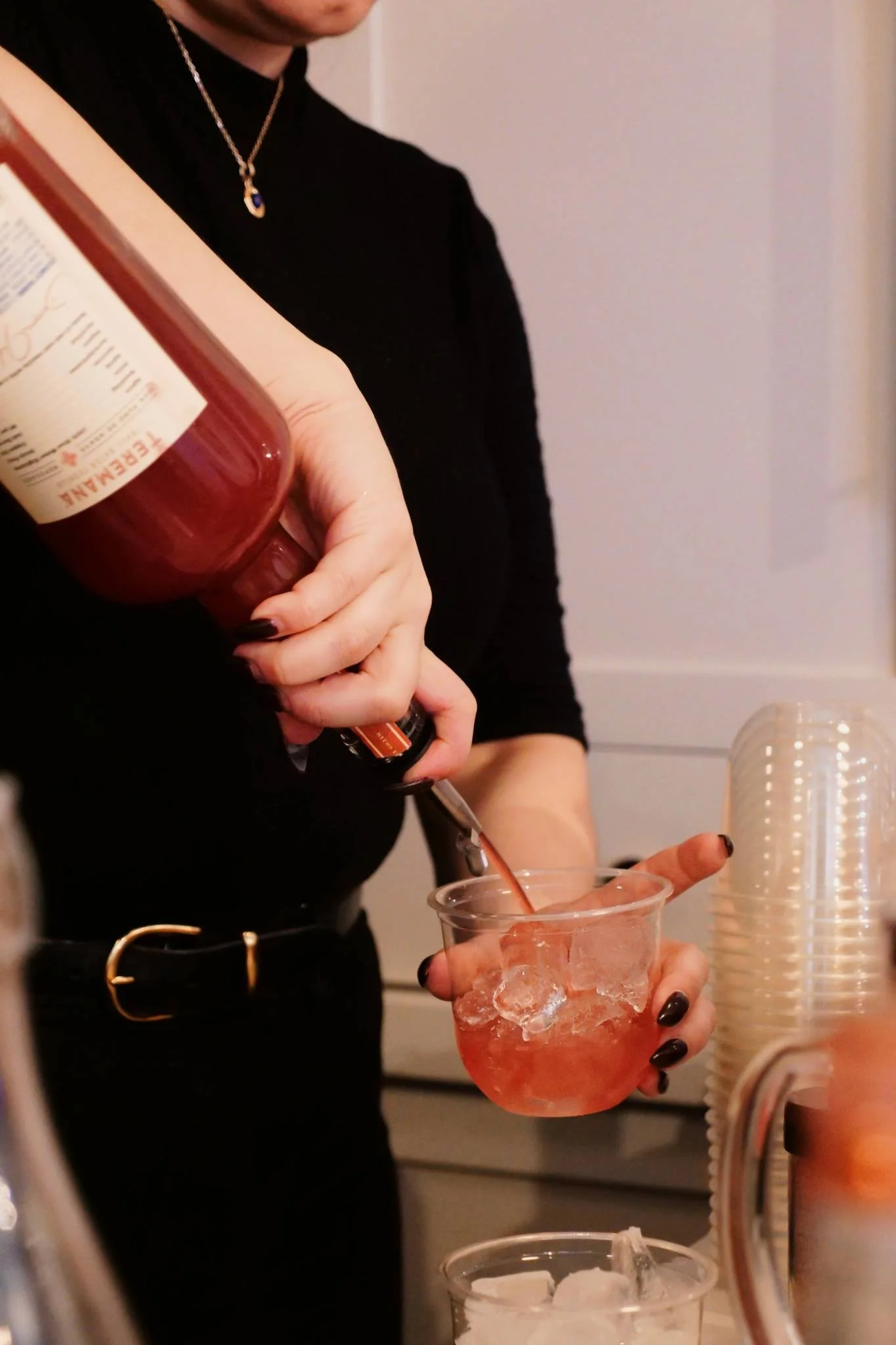 Person pouring red liquid into a clear plastic cup with ice, wearing a black top and necklace, with a stack of paper cups nearby.