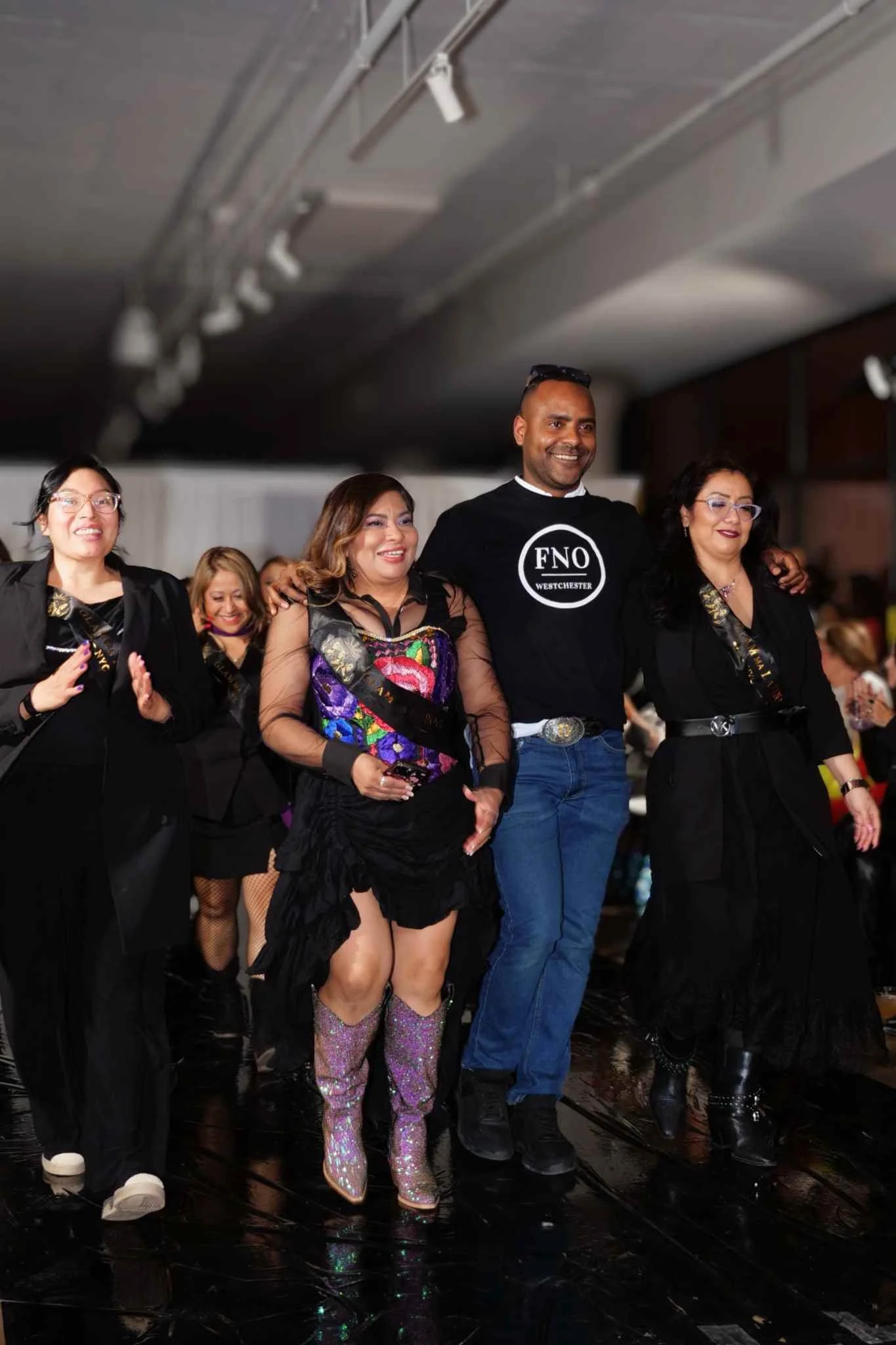 Group of people, including a woman in shiny purple boots and a woman in black with a colorful top, standing together in a line at a social event, smiling.