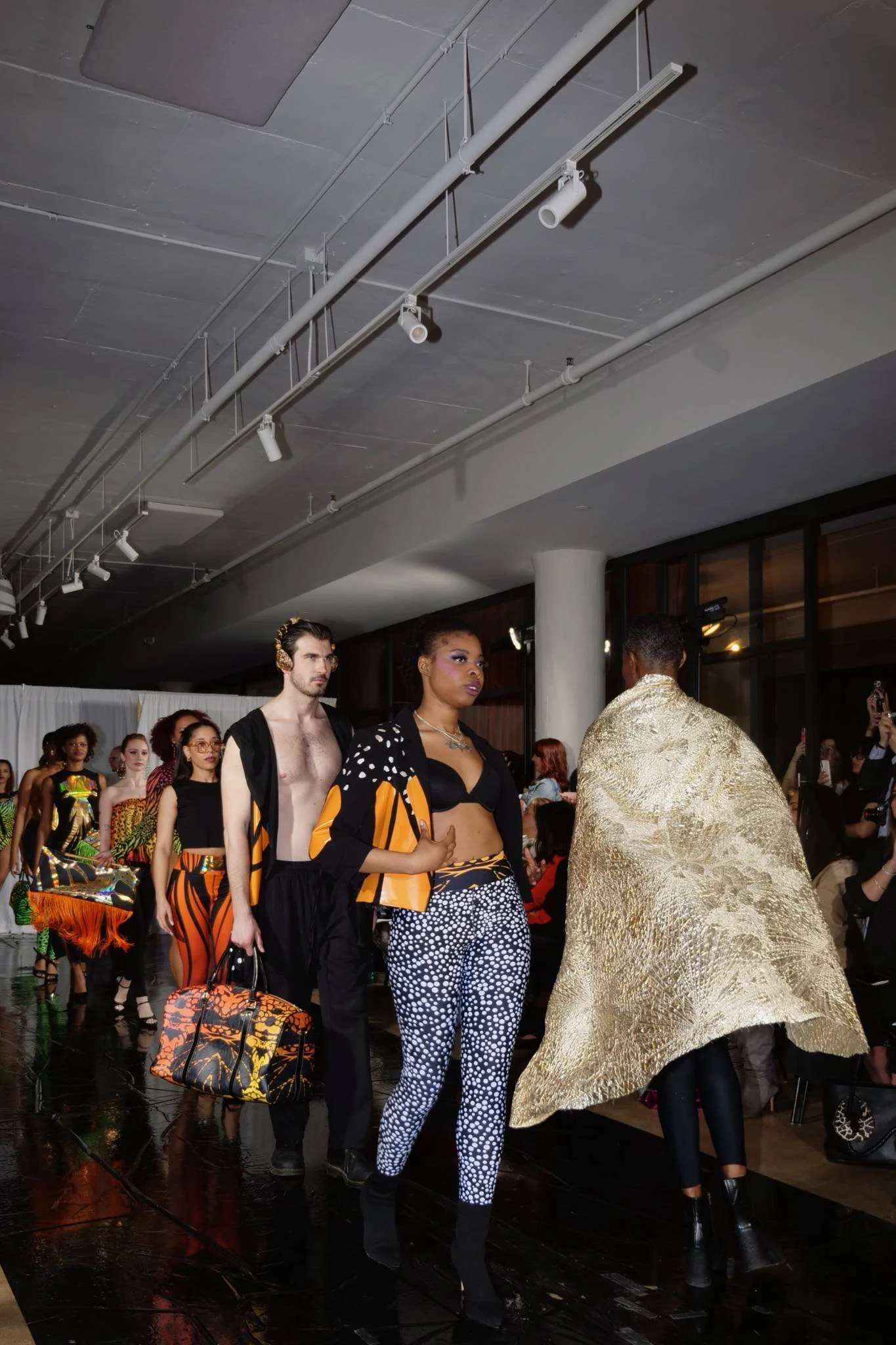 Models walk in a fashion show wearing vibrant and eclectic clothing, with a diverse group of spectators watching indoors under ceiling track lighting.