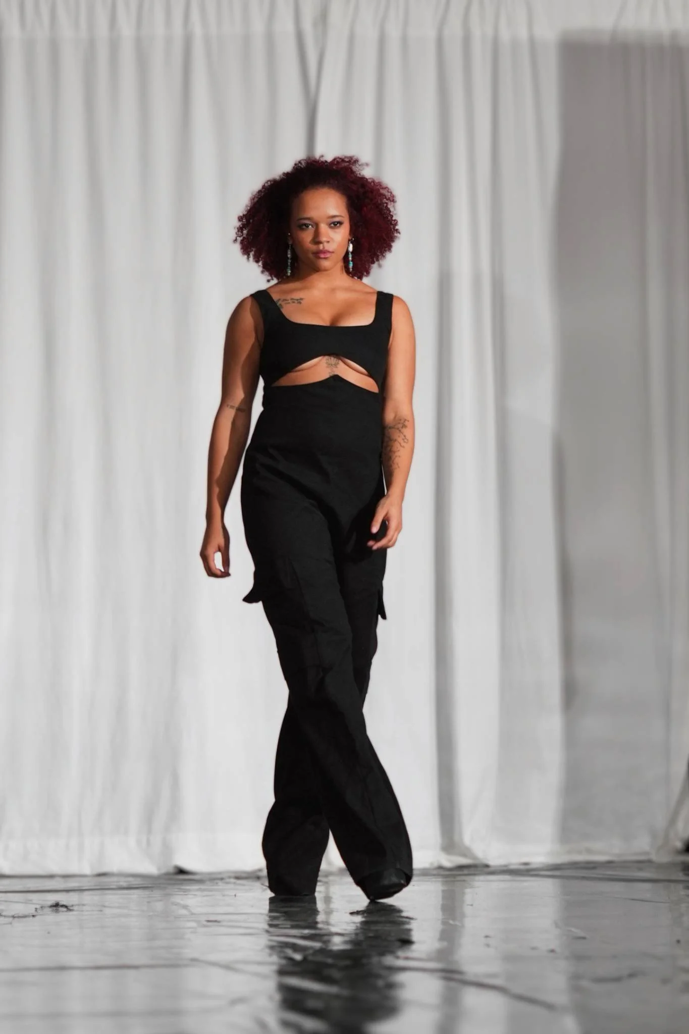Woman with curly red hair walking across a stage with a white curtain background, dressed in a black outfit.
