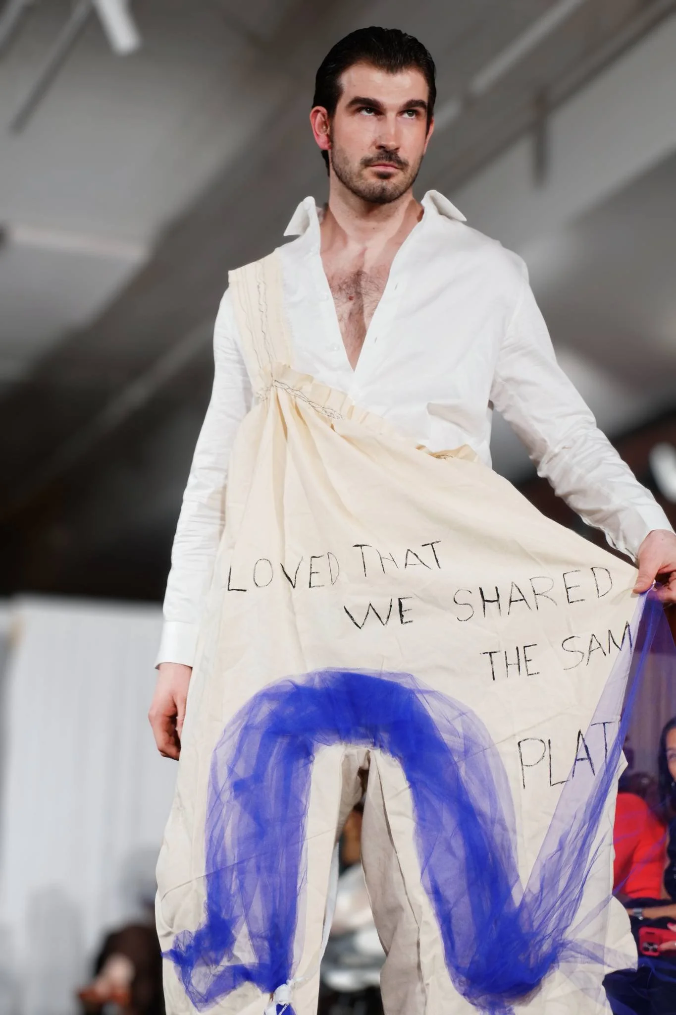 A man in an off-white shirt and baggy pants with blue painter's tape or paint on them walking in an indoor space, with a woman in a black and red outfit looking at him in the background.